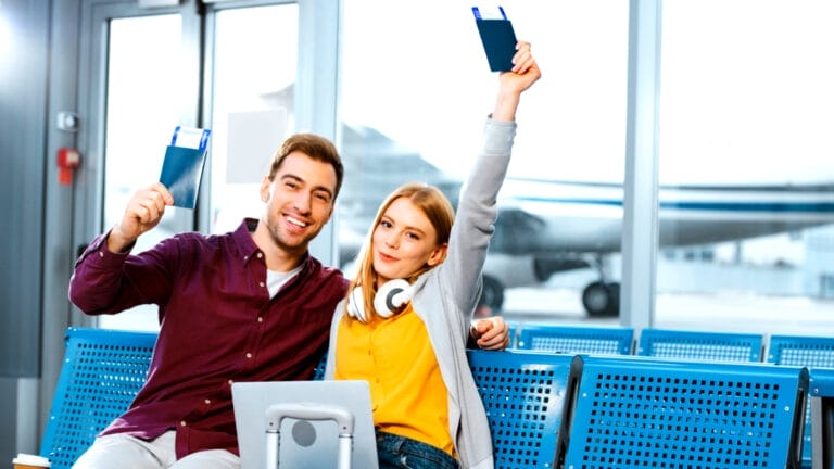 Couple sitting in an airport holding their passports with tickets in the air.